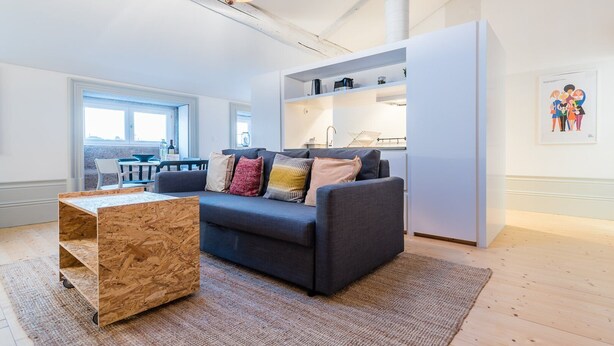A modern living room with a dark grey sofa, a wooden coffee table, and a kitchen area visible through an open door. - GuestReady - Mezzanine at the Bridge (Porto)
