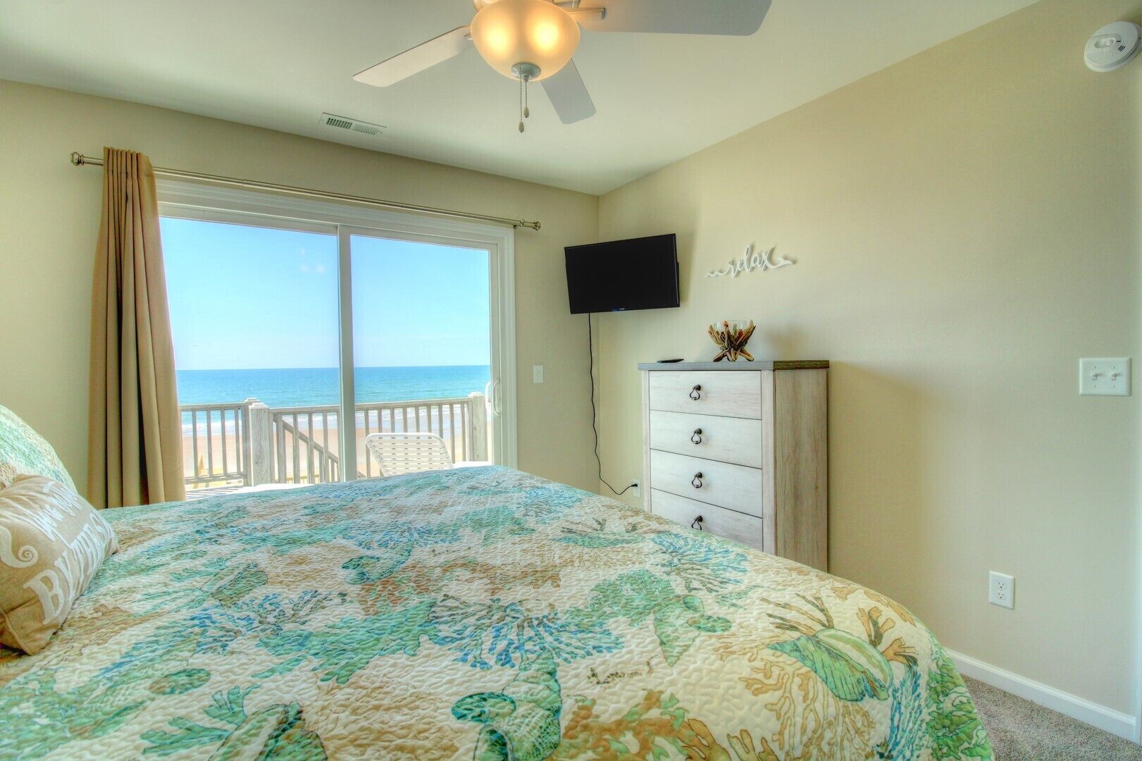 A bedroom with a large bed, a ceiling fan, a TV, and a view of the beach.
