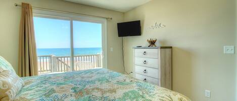 A bedroom with a large bed, a ceiling fan, a TV, and a view of the beach.