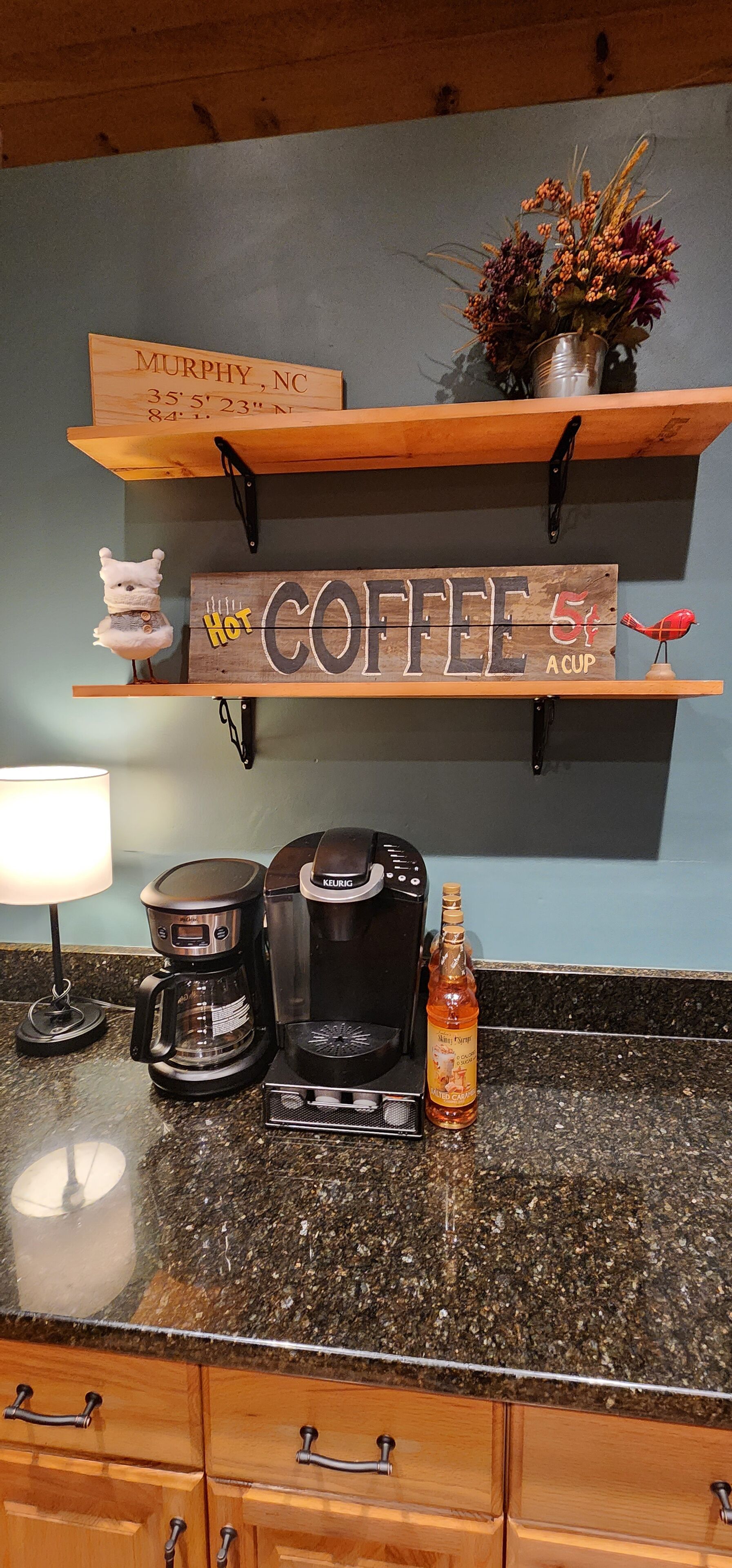 A kitchen counter with a coffee maker, a Keurig, a mug, and a bottle of syrup. Above the counter, there are wooden shelves with a sign that says "Coffee 5¢ a cup" and a small decorative item.
