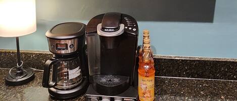 A kitchen counter with a coffee maker, a Keurig, a mug, and a bottle of syrup. Above the counter, there are wooden shelves with a sign that says "Coffee 5¢ a cup" and a small decorative item.