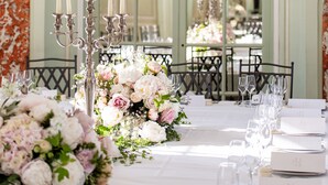 A formal dining table set with floral centerpieces, candle holders, and place cards, with a view of a green arched window and a chandelier in the background. - Château with Spa, Golf, Tennis, Basket, swimming pools, gardens.  (Villers-Bocage)