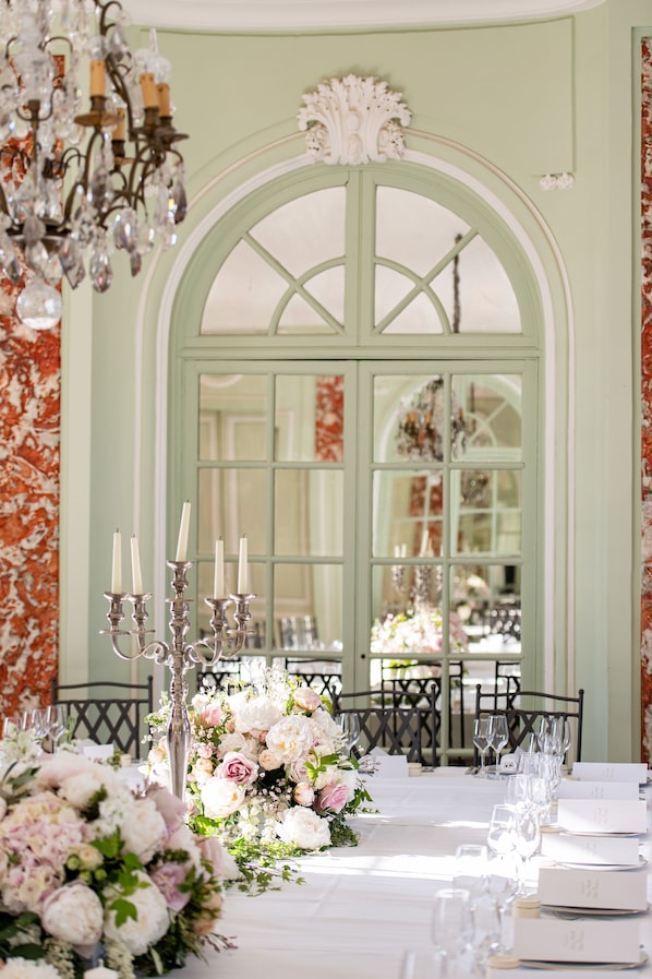 A formal dining table set with floral centerpieces, candle holders, and place cards, with a view of a green arched window and a chandelier in the background.