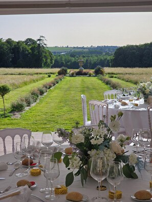 An outdoor dining area with a table set for a meal, surrounded by chairs, and a view of a manicured garden and fields. - Château with Spa, Golf, Tennis, Basket, swimming pools, gardens.  (Villers-Bocage)
