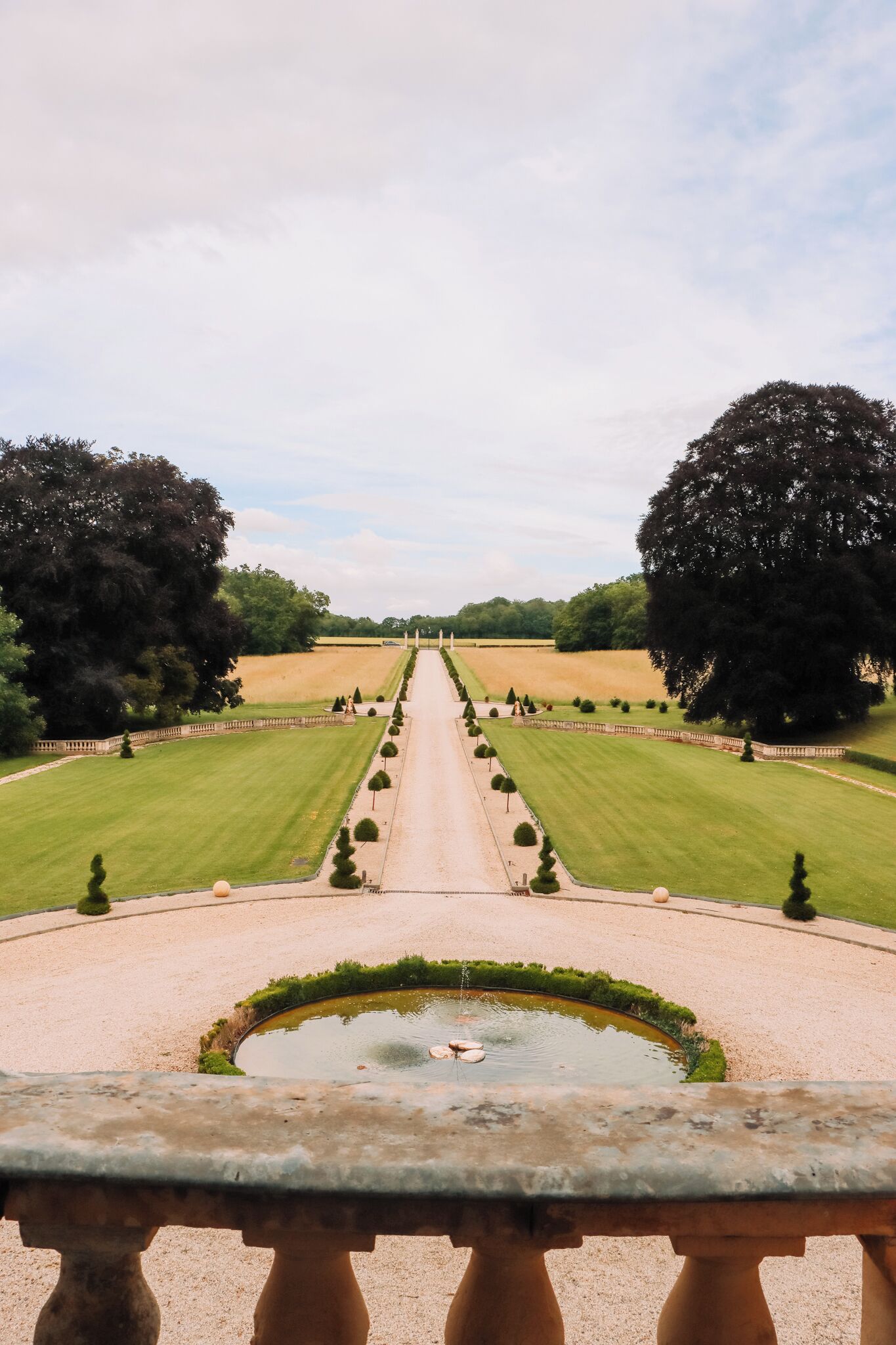 Um jardim formal com um caminho central que leva a um lago, rodeado por sebes e árvores bem aparadas.