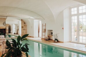 A spacious indoor swimming pool area with a clear view of the pool, a lounge area with seating, and a stone wall. - Château with Spa, Golf, Tennis, Basket, swimming pools, gardens.  (Villers-Bocage)
