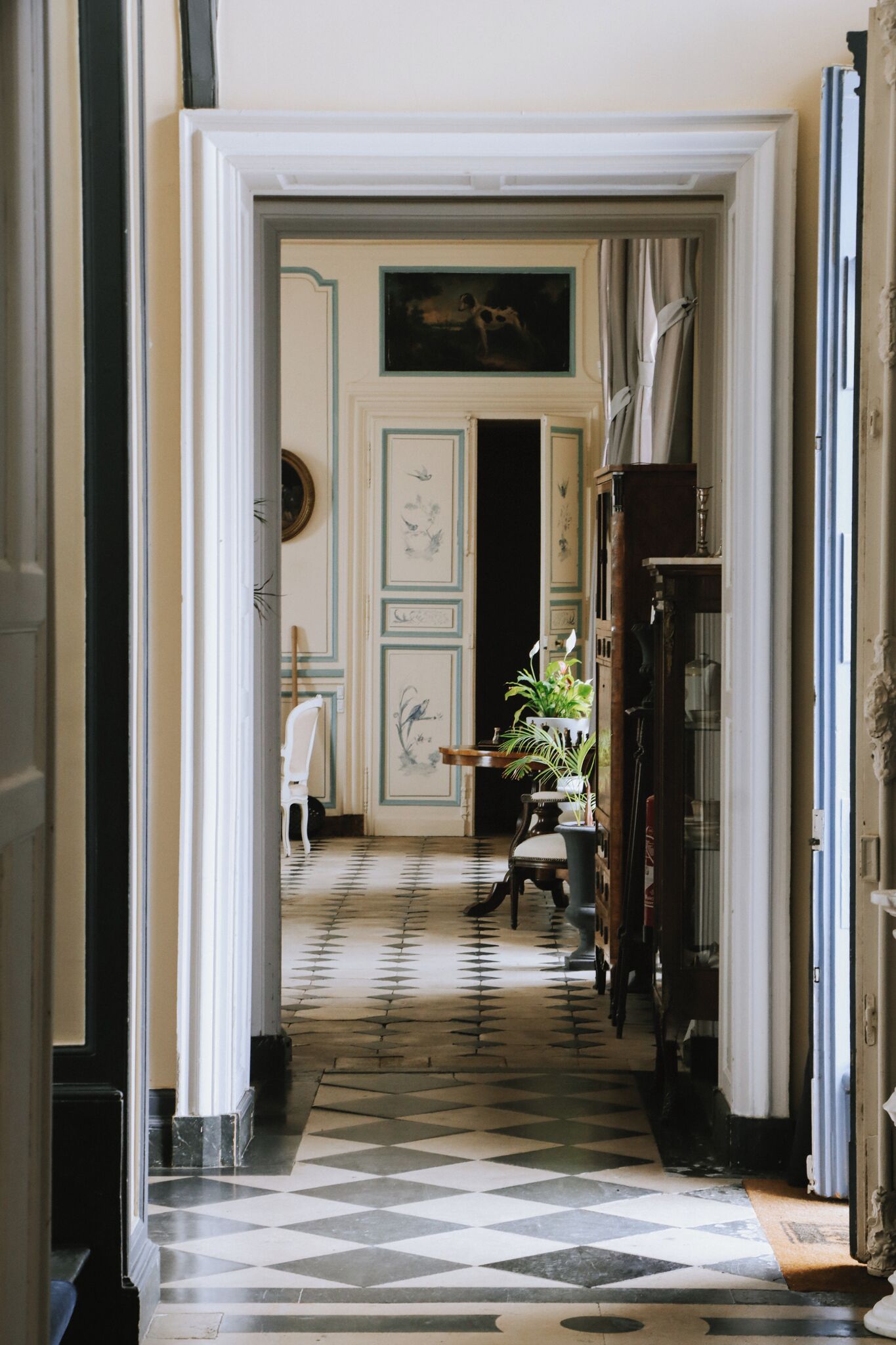 A hallway with a checkered floor, a white chair, a table with plants, and a painting on the wall.