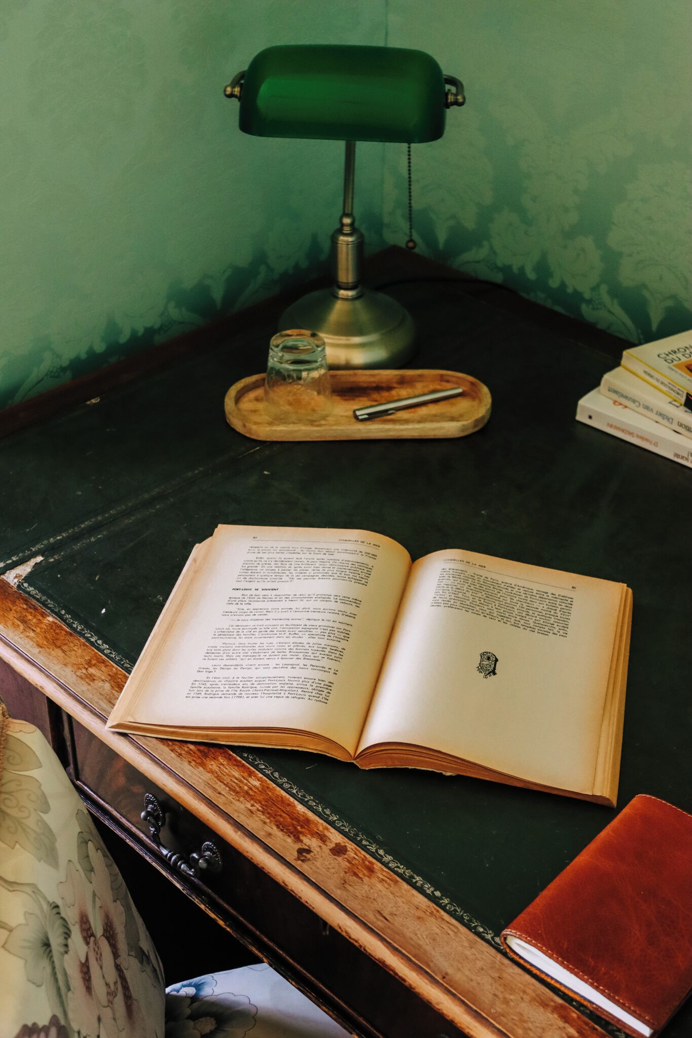An open book on a desk with a green desk lamp, a glass, and a pen.
