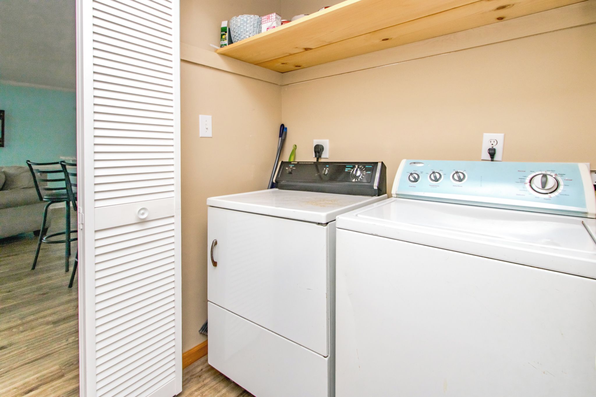 A laundry room with a front-loading washer and dryer, a shelf above the dryer, and a door leading to a living area.