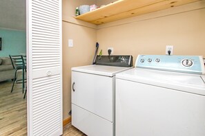 A laundry room with a front-loading washer and dryer, a shelf above the dryer, and a door leading to a living area.