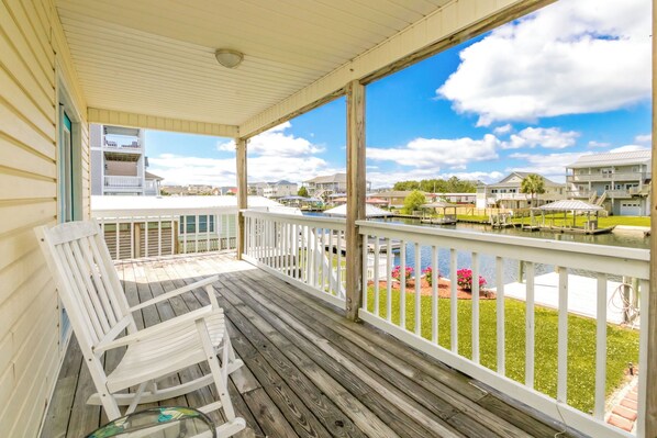 A wooden deck with white railings, a white rocking chair, and a view of a marina with boats and houses in the background. - Mama's Pride - 3BR Home on the Canal - Boat Dock & Sound Views! (Surf City)