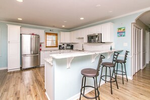 A modern kitchen with white cabinetry, stainless steel appliances, and a central island with bar stools. - Mama's Pride - 3BR Home on the Canal - Boat Dock & Sound Views! (Surf City)