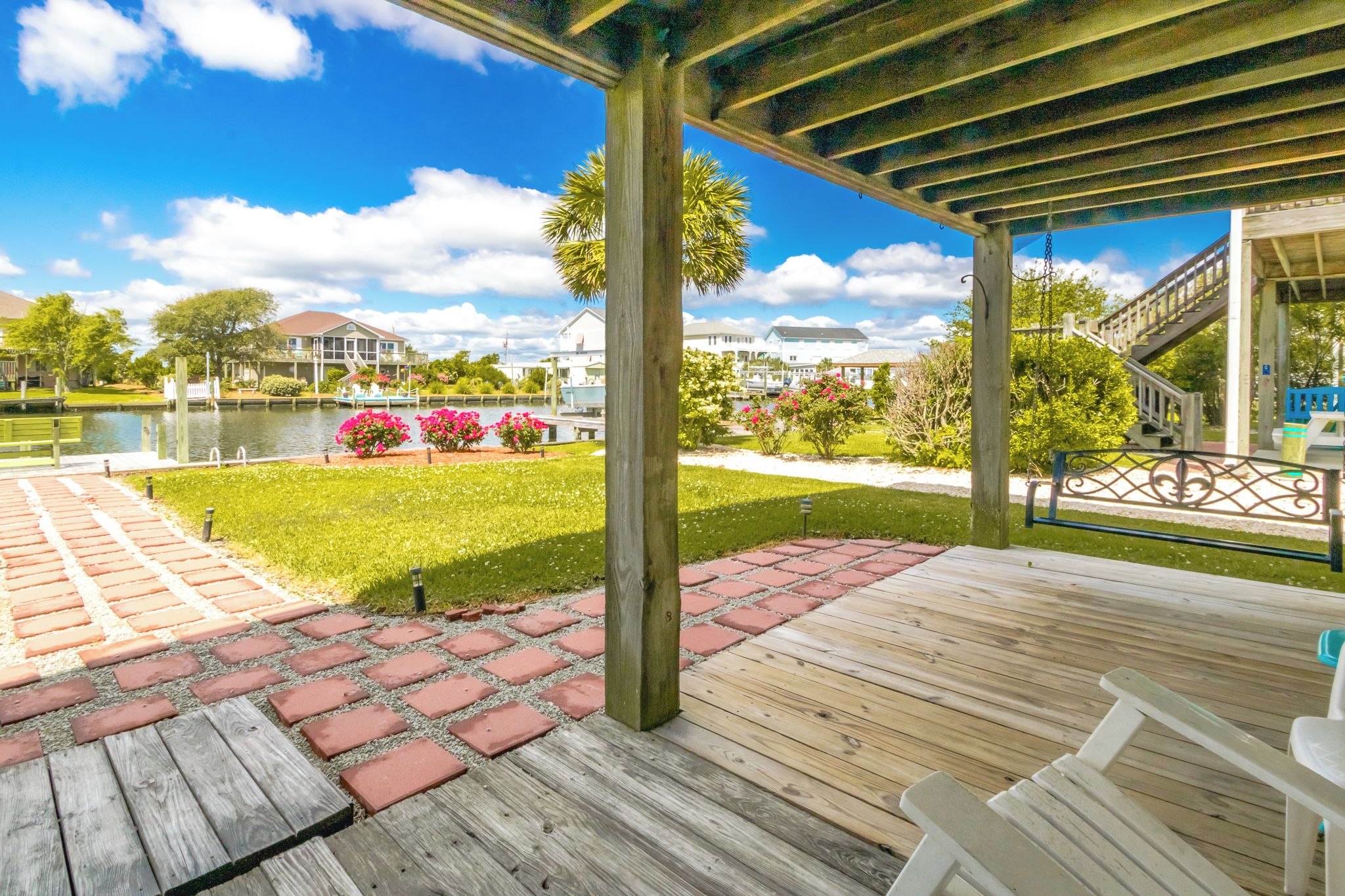 A covered outdoor area with a wooden deck, a brick pathway, and a view of a lake with houses in the background.