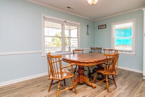 A dining room with a round wooden table and six chairs, large windows with blinds, and a light blue wall.