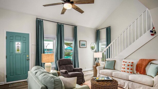 A living room with a teal door, a ceiling fan, a white staircase, and a white sofa with orange pillows. - Dream Weaver: Spacious 4BR home in Holden Beach, perfect for family getaways! (Holden Beach)