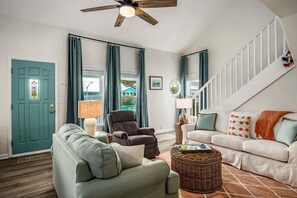 Una sala de estar con una puerta verde azulada, un ventilador de techo, una escalera blanca y un sofá blanco con almohadones naranja.