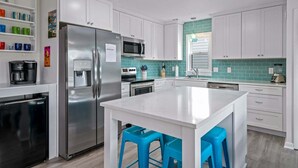 A modern kitchen with white cabinets, a stainless steel refrigerator, a microwave, and a teal backsplash. - Dream Weaver: Spacious 4BR home in Holden Beach, perfect for family getaways! (Holden Beach)