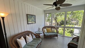 Une véranda couverte, meublée de meubles en osier, dotée d’un ventilateur de plafond et offrant une vue sur la verdure environnante.