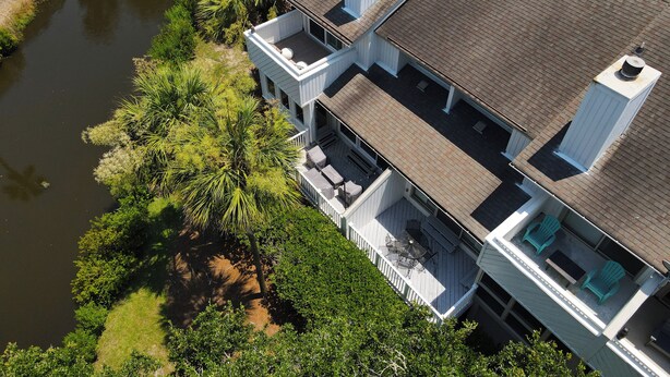 Une maison à deux étages dotée d’un balcon et d’un porche abrité, entourée d’une végétation luxuriante et bordée par une rivière.