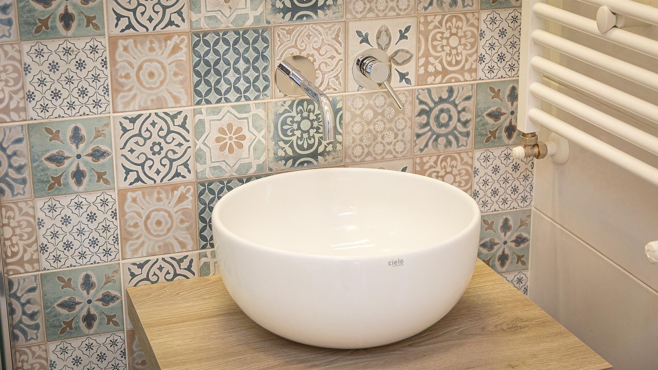 A bathroom with a white ceramic sink on a wooden platform, surrounded by a mosaic tile wall.