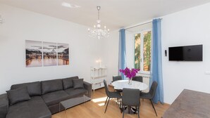 Una sala de estar moderna con un sofá seccional gris, una mesa de comedor redonda con sillas, una lámpara de araña, un televisor de pantalla plana montado en la pared y una ventana con cortinas azules.