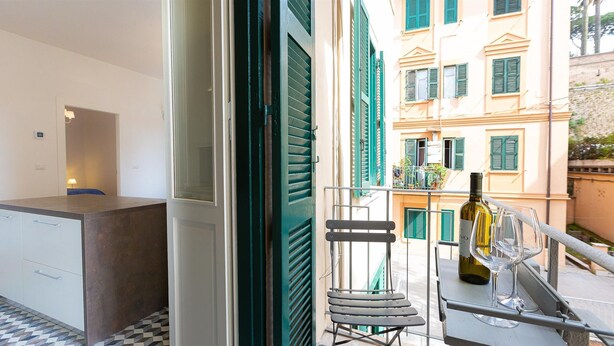 Habitación con vistas a un edificio con contraventanas verdes, una mesa con botella de vino y copas, y una silla.