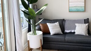 A modern living room with a grey sofa, a large potted plant, and a beach-themed artwork on the wall.