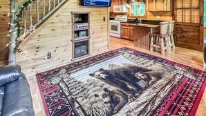 A cozy cabin interior with a wooden staircase, a flat-screen TV, a kitchen area, and a large rug with a bear design.