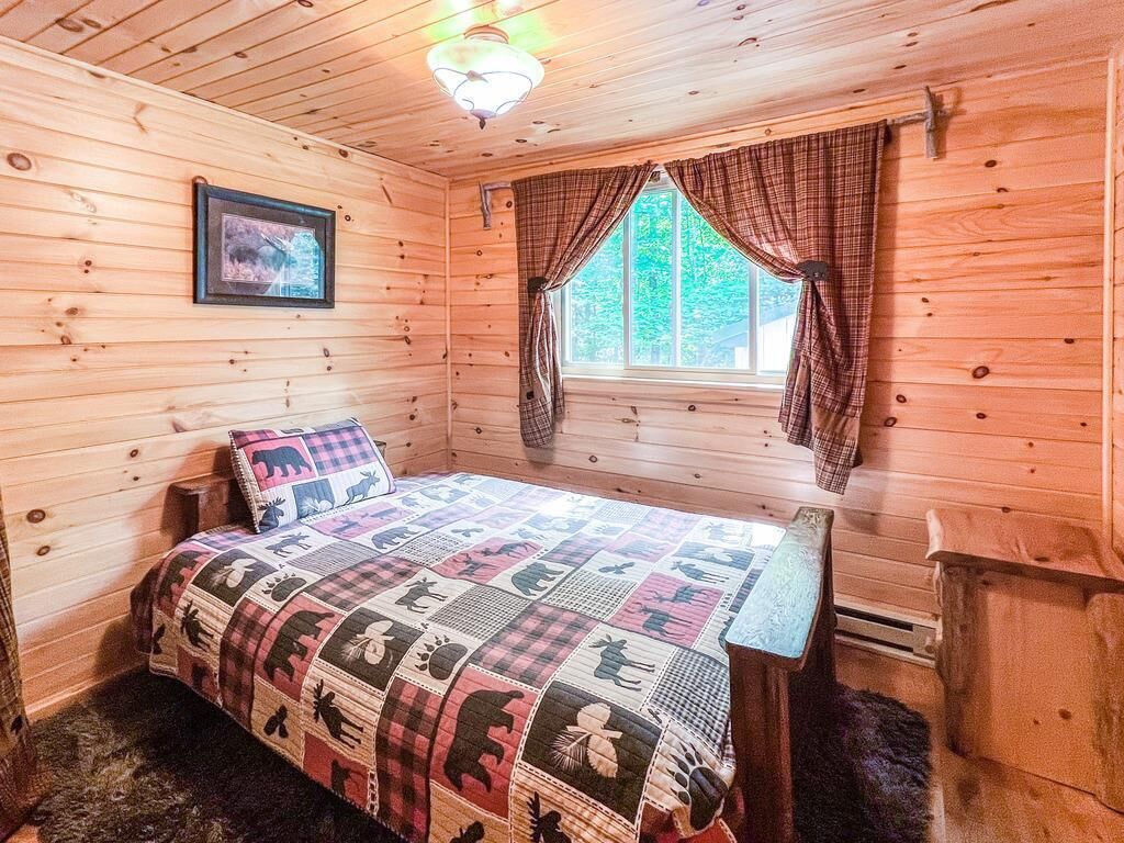 A wooden cabin room with a bed, a window with curtains, a framed picture on the wall, and a small table.