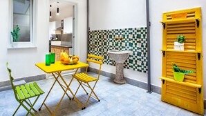 Une cuisine de taille modeste, avec une table jaune, des chaises pliantes vertes, un crédence à carreaux et une porte jaune ornée d’un pot de fleurs.