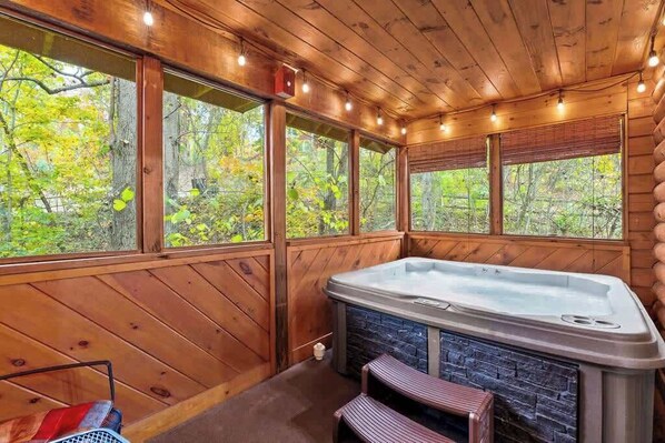 A wooden cabin interior with a hot tub, large windows, and a stone fireplace.