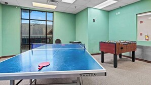 A room with a ping pong table, a foosball table, and a window with a view of a building. - Cozy Mountain Retreat w Pools &Free Attraction Tix (Pigeon Forge)