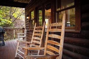 A wooden rocking chair on a wooden deck with a log cabin in the background.