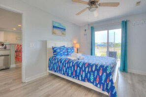 A bedroom with a bed, a ceiling fan, a view of the beach, and a balcony with curtains. - OW 9101 - Days Drift By: A cozy oceanview condo in Oak Island, perfect for families. (Oak Island)