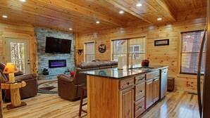 A cabin interior with a stone fireplace, wooden floors, a kitchen island, and a living area with a sofa and TV. - Close To Pigeon Forge Attractions/ Seasonal Outdoor Pool/ Perfect For Families! (Pigeon Forge)