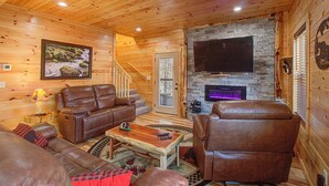 A log cabin interior with a fireplace, leather furniture, and a mounted TV. - Close To Pigeon Forge Attractions/ Seasonal Outdoor Pool/ Perfect For Families! (Pigeon Forge)