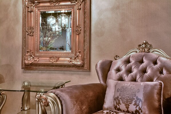 A luxurious room with a tufted velvet armchair, a glass coffee table, and an ornate mirror with a floral frame.
