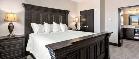 Un dormitorio con una cama grande, dos mesitas de noche con lámparas, un cabecero de madera oscura y una puerta que da al baño.