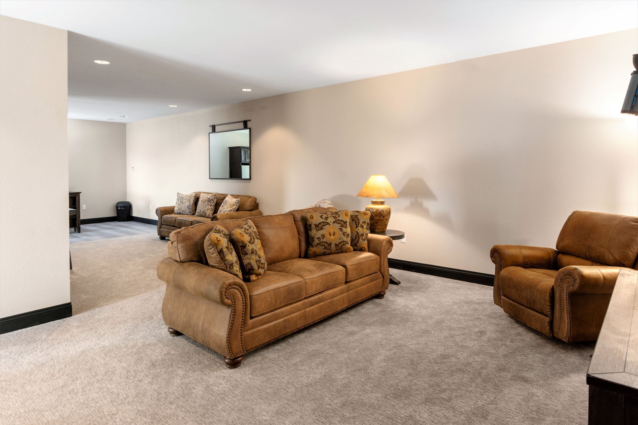 A living room with a brown leather sofa, a matching armchair, a side table with a lamp, and a large mirror on the wall.