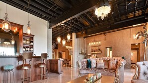 A spacious, elegantly furnished living area with a high ceiling and exposed beams, featuring a bar area with a wine rack and stools, a plush sofa set, and ornate lighting fixtures. - Hotel 21 & Co. (Gering)