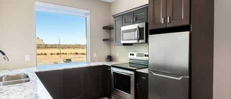 A modern kitchen with dark cabinetry, stainless steel appliances, and a large window offering a view of a desert landscape.
