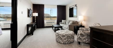 A hotel room with a sofa, ottoman, and a small table with a lamp. There is a TV mounted on the wall and a window with curtains.