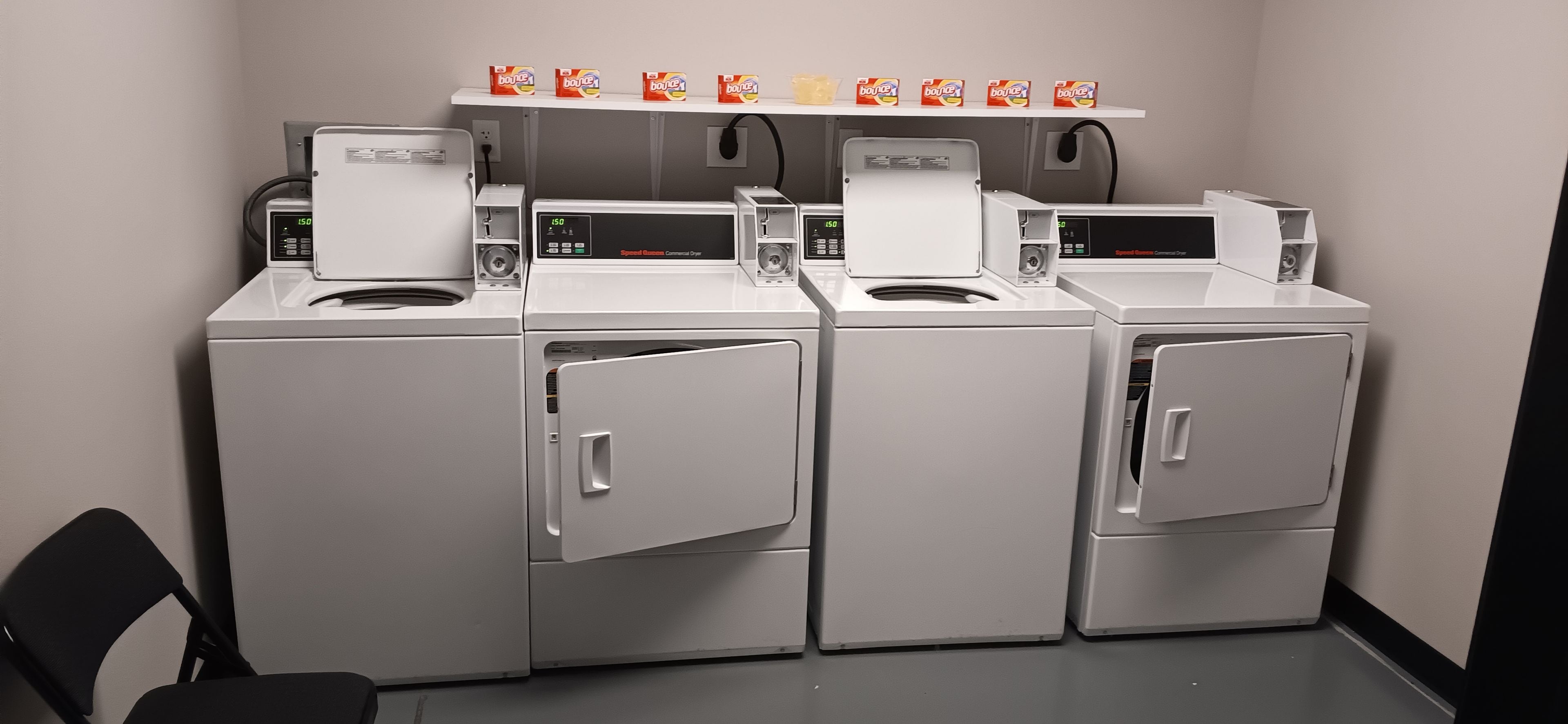 Een wasserette met vier wasmachines en drogers, een plank met wasmiddel en een stoel.