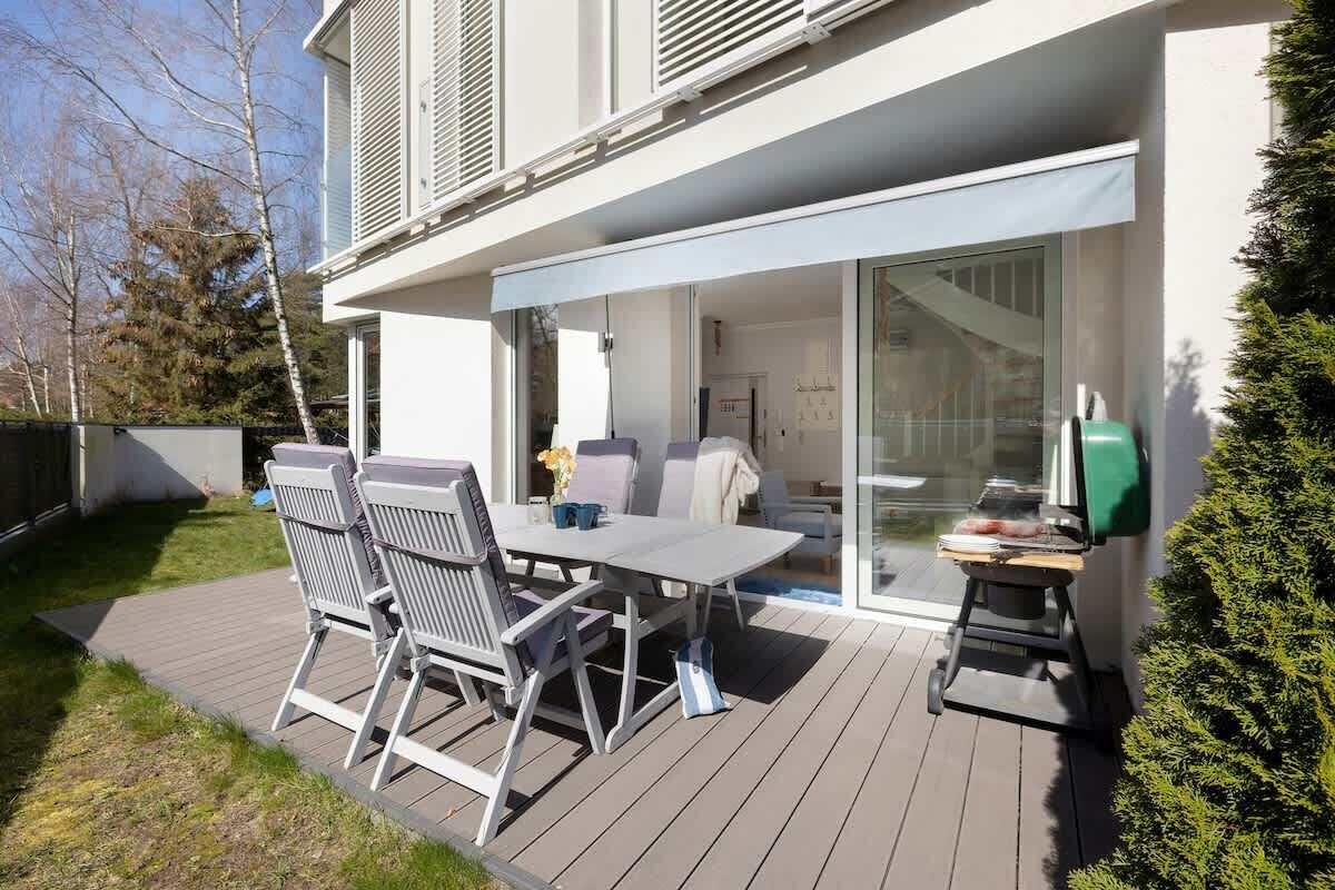 A well-furnished terrace with a table, chairs, and a parasol, surrounded by a green garden.