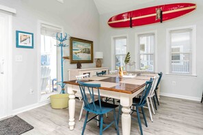 Un coin repas avec une table en bois et des chaises bleues, une planche de surf rouge accrochée au mur et un tableau représentant une scène de plage.