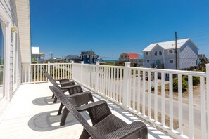 Un balcon avec des fauteuils en osier et une balustrade blanche, donnant sur une plage bordée de maisons.