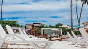 Een strand met witte ligbedden en parasols, palmbomen en een houten constructie op de achtergrond.