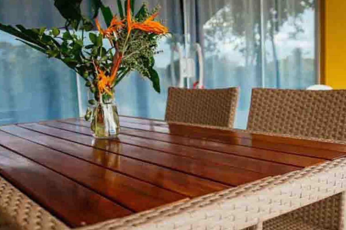 A wooden dining table with woven chairs, a vase with flowers, and a view of a yellow building through large windows.