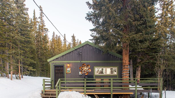 Una cabaña con fachada verde, una terraza de madera y suelo cubierto de nieve.