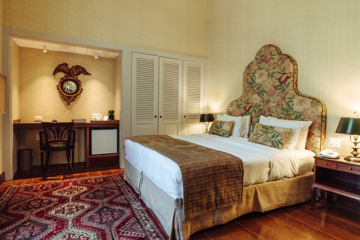 A bedroom with a large bed, a desk, and a chair. There is a mirror on the wall and a door with louvered shutters.